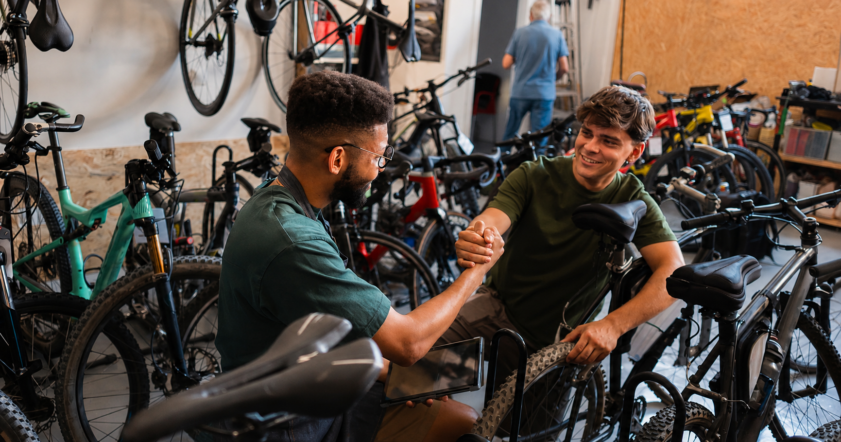 A bicycle shop employee and a customer smile as they shake hands beside a mountain bike in a busy showroom filled with bicycles. Additional bikes hang on the walls and line the floor, while another person works in the background, creating a welcoming, community-focused atmosphere.