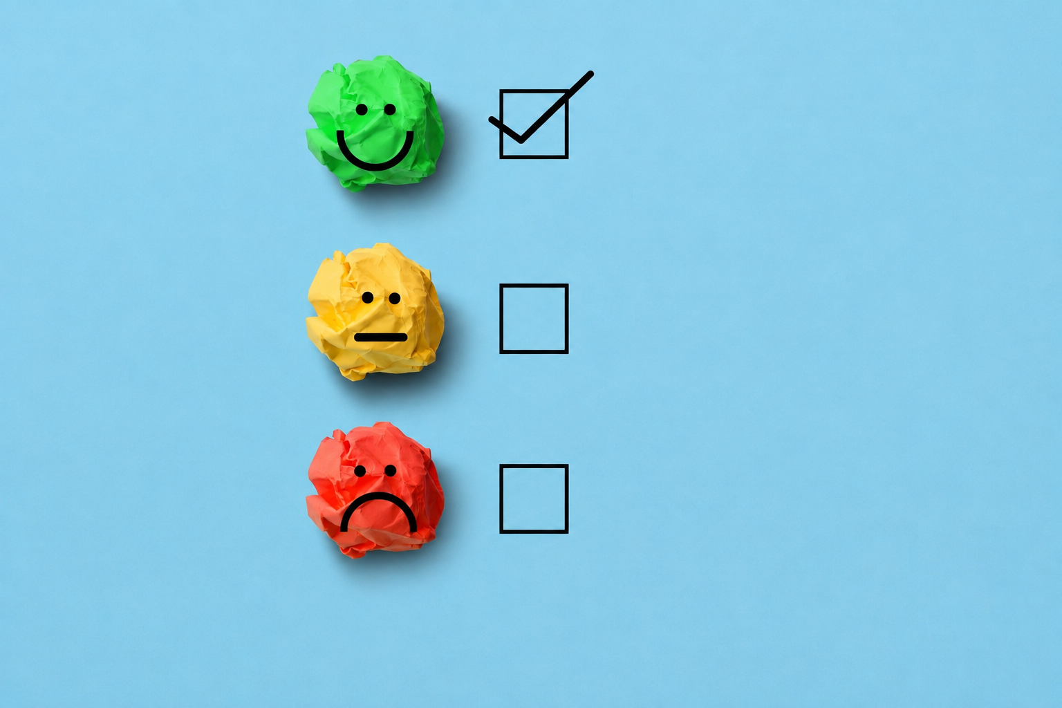 Three crumpled paper balls with hand-drawn faces represent customer satisfaction levels against a light blue background. A green smiling face at the top has a checked box beside it, while a yellow neutral face and a red sad face below each have empty checkboxes, illustrating a positive customer feedback or satisfaction survey concept.