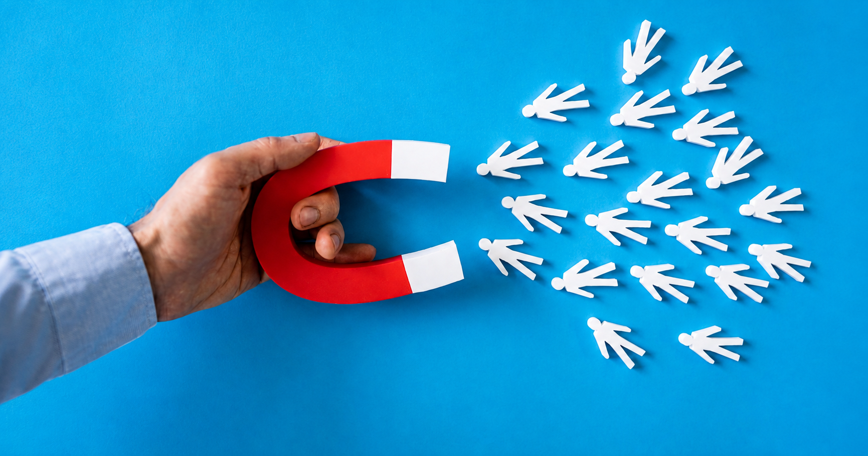 A hand holds a red horseshoe magnet with white tips against a bright blue background, attracting a scattered group of small white person-shaped icons clustered toward the magnet. The image symbolizes attracting people, customers, talent, or leads.