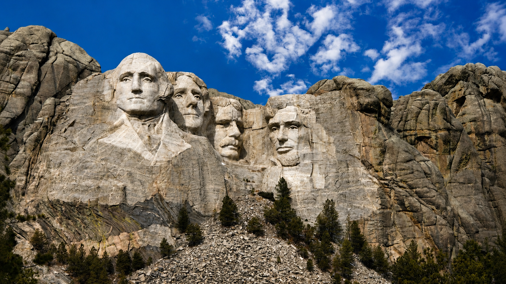 Mount Rushmore National Memorial carved into a granite mountainside under a bright blue sky with scattered white clouds. The monumental faces of George Washington, Thomas Jefferson, Theodore Roosevelt, and Abraham Lincoln are prominently featured in the rock formation, surrounded by rugged cliffs and outcrops. A slope of loose stone and evergreen trees fills the foreground, emphasizing the scale of the monument and the natural landscape of South Dakota&rsquo;s Black Hills.